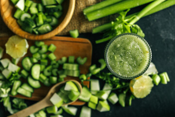 Fresh green smoothie in a textured glass surrounded by chopped ingredients including cucumber,...