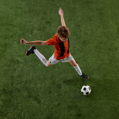 Top view image of boy, soccer player in orange jersey and white shorts in motion of green field,...