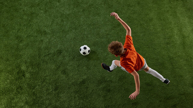Image viewed from above of boy, soccer player in orange jersey and white shorts in motion of green field, playing, dribbling ball. Training activity. Concept of sport, education, competition, game