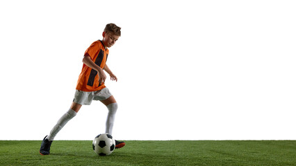 Focused boy, soccer player in orange jersey and white shorts training, dribbling ball on grassy surface against white studio background. Concept of sport, education, competition, game