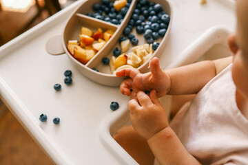 Toddler self-feeding with sliced peaches, bananas, and blueberries from a divided plate on a highchair. Perfect for baby-led weaning, healthy snacking, and developing fine motor skills