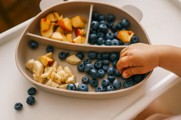 Toddler self-feeding with sliced peaches, bananas, and blueberries from a divided plate on a highchair. Perfect for baby-led weaning, healthy snacking, and developing fine motor skills
