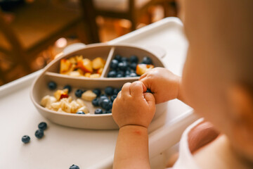 Toddler self-feeding with sliced peaches, bananas, and blueberries from a divided plate on a highchair. Perfect for baby-led weaning, healthy snacking, and developing fine motor skills