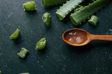 Fresh aloe vera gel in a wooden spoon with cut aloe leaves on a dark textured surface. Natural...