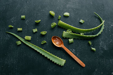 Fresh aloe vera gel in a wooden spoon with cut aloe leaves on a dark textured surface. Natural...