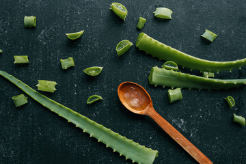 Fresh aloe vera gel in a wooden spoon with cut aloe leaves on a dark textured surface. Natural...