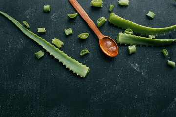 Fresh aloe vera gel in a wooden spoon with cut aloe leaves on a dark textured surface. Natural...