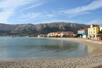 A serene late autumn scene of Vela Plaza (Great Beach) in Baska, Croatia, featuring a calm sea against a backdrop of a coastal village and rocky mountains