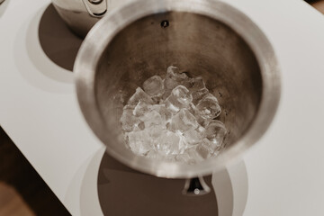  A close-up top-down view of a stainless steel ice bucket filled with partially melted ice cubes, placed on a white countertop with another metal container in the background