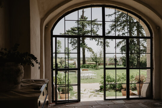 A picturesque view of a lush green garden with an outdoor wedding ceremony setup, framed by an elegant arched glass door with black metal framing