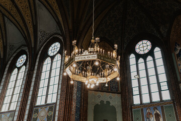 An ornate chandelier with glowing candles and intricate metalwork hangs from the high vaulted ceiling of a historic church, surrounded by towering stained glass windows