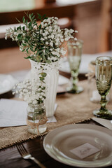 Elegant rustic wedding table setting with a burlap table runner, delicate white baby's breath flowers arranged in a ceramic vase and glass jar tied with twine, complemented by vintage green glass 