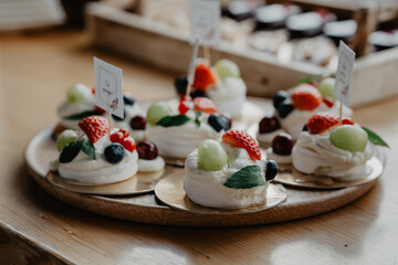 A beautifully arranged wooden serving platter featuring delicate pavlova desserts topped with fresh fruits such as strawberries, grapes, blueberries, and red currants, garnished with mint leaves