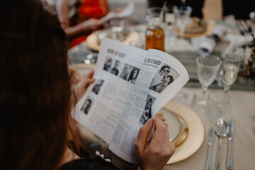 A guest at an elegantly set dining table attentively reads a black-and-white printed wedding newspaper featuring vintage photographs and articles, surrounded by fine glassware, gold-rimmed plates