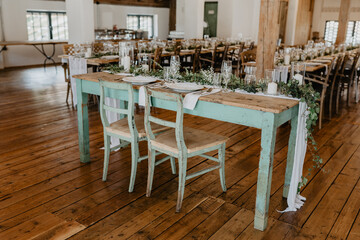 A beautifully arranged rustic wedding sweetheart table with vintage turquoise wooden chairs, elegant white tableware, polished silverware, and a delicate floral greenery centerpiece intertwined 