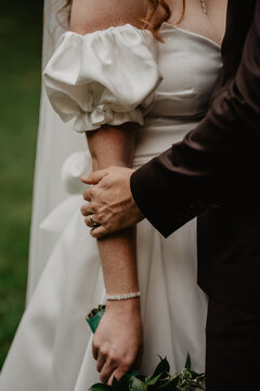  A groom in a dark suit tenderly holding the bride&rsquo;s arm, showcasing their wedding rings, as she wears a white satin dress with elegant puff sleeves, a delicate diamond bracelet, and holds a bouquet 