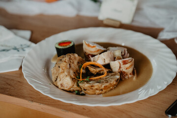  A gourmet plated dish featuring bacon-wrapped chicken medallions, traditional bread dumplings, rich brown sauce, and a decorative vegetable garnish, served on a white porcelain plate 