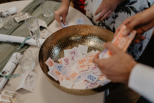 A close-up of a golden raffle bowl filled with colorful numbered tickets at an elegant event, where guests' hands are seen drawing and organizing tickets on a beautifully decorated table 