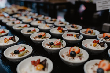  A beautifully arranged dessert buffet featuring multiple small white bowls filled with creamy dessert topped with fresh strawberries, blueberries, kiwi, chocolate pieces, and golden berries, elegantl
