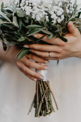  A bride's delicate hands with a simple diamond wedding ring gently holding a lush bridal bouquet made of white baby's breath flowers and green olive leaves, wrapped in white ribbon with natural woode