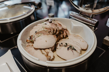 A close-up of a buffet station featuring a serving dish filled with tender, sliced roasted pork garnished with microgreens, presented in a professional catering setup with stainless steel 