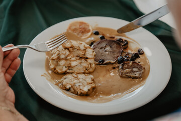 A traditional Czech dish featuring sliced roasted meat with a creamy sauce, bread dumplings, and garnished with cranberries and a slice of lemon served on a white plate with cutlery on a green tablecl