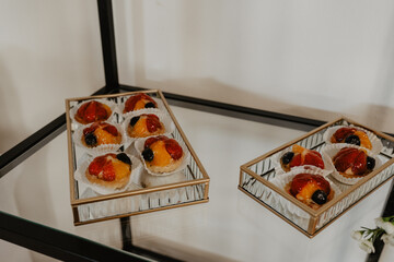  Elegant fruit tarts with a glossy glaze, featuring fresh strawberries, blueberries, and orange slices, beautifully presented in gold-trimmed glass trays on a reflective dessert stand