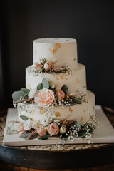 A luxurious three-tier wedding cake adorned with fresh pastel roses, baby's breath flowers, and delicate gold leaf accents, placed on a rustic wooden barrel, creating an elegant and romantic wedding d