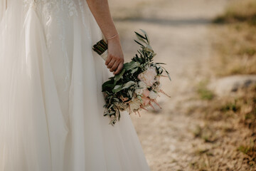 A bride in a flowing white wedding dress holding a romantic bouquet of soft pastel flowers and greenery while walking along a sunlit countryside path, capturing an elegant and natural moment 
