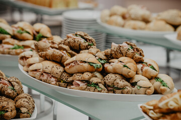 A beautifully arranged platter of freshly made croissant sandwiches filled with ham, lettuce, and cheese, served at a catering event with a selection of seeded and plain bread rolls in the background