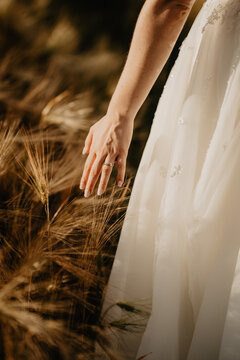  A close-up of a bride&rsquo;s hand gently brushing against golden wheat stalks as she walks through a sunlit field, wearing an elegant wedding dress with delicate embroidery and a gold wedding 