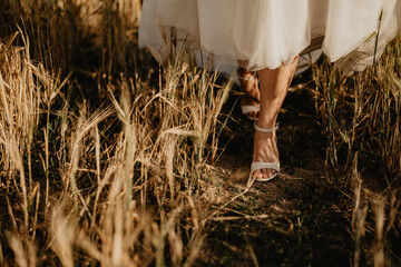 A romantic close-up of a bride walking gracefully through a golden wheat field in soft evening light, with the flowing hem of her delicate wedding dress brushing against the tall stalks as she steps 