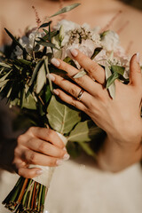  A close-up of a bride’s hands holding a lush wedding bouquet of green leaves and delicate white flowers, showcasing her elegant engagement ring and wedding band along with beautifully manicured nails