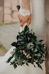 A close-up of a bride's hand delicately holding a rustic wedding bouquet made of lush green eucalyptus leaves and small white flowers, wrapped in natural lace fabric