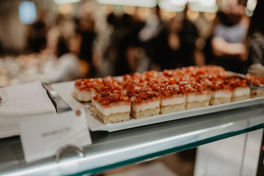 A tray of beautifully arranged layered dessert bars topped with fresh diced strawberries and glaze displayed on a glass counter at a catering event with blurred guests in the background