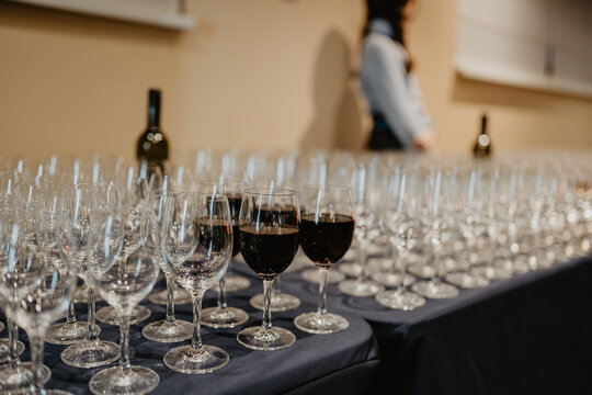 A table covered with a dark blue cloth set up for a formal event featuring neatly arranged empty wine glasses with a few filled with red wine in the foreground and a blurred bartender standing in the 