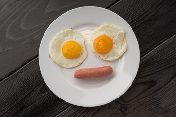 A white plate with two fried eggs and a single sausage on a dark wooden table.