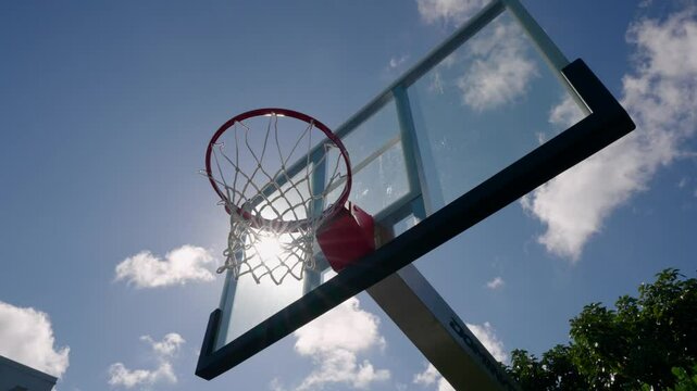 Basketball hoop with backboard against bright blue sky with some clouds. Sun is shining brightly creating lens flare effect. Basketball hoop seen from low angle