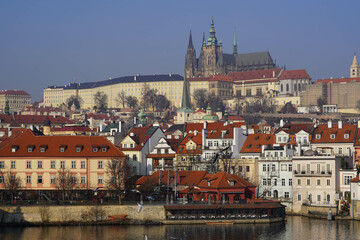 Obraz premium The cityscape shows colorful buildings with red rooftops near a river, with Prague Castle towering in the background