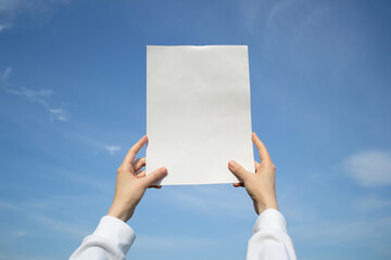 Hands holding up a blank sheet of paper outdoors, on a blue sky background.