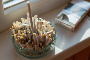 A dirty ashtray full of cigarette butts against a window background. 
