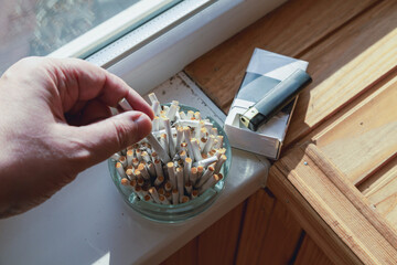 A smoker's hand extinguishing an unfinished cigarette in an ashtray overflowing with butts on a...