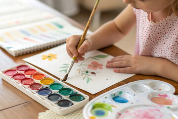 Naklejka premium A Child's Imagination: A young girl, immersed in her creativity, paints a beautiful flower on a sheet of paper using vibrant watercolor paints.