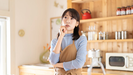 キッチンで考えごとをするエプロン姿の女性
