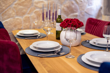 Elegant dining table set with white porcelain plates, wine glasses, and silverware, featuring a red rose centerpiece in a stylish white vase
