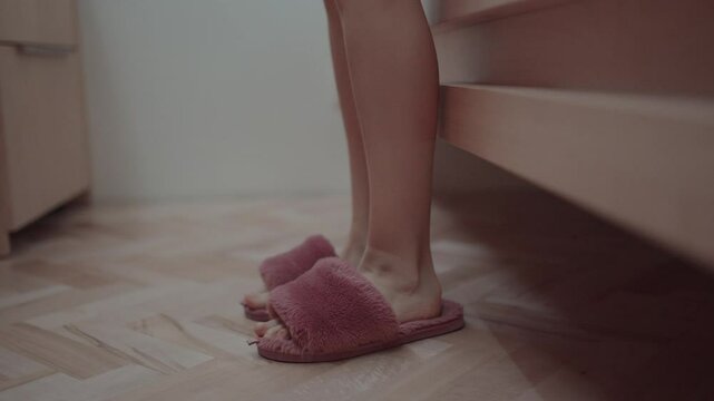 A person&rsquo;s bare feet stepping into soft pink slippers beside a bed on a wooden floor, capturing a cozy and relaxing morning moment.
