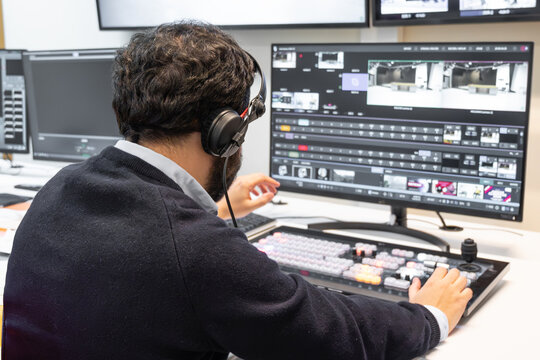 A television director in a broadcast control room, actively guiding a live production. Surrounded by multiple screens, the director wears a headset and gestures towards a video switcher.