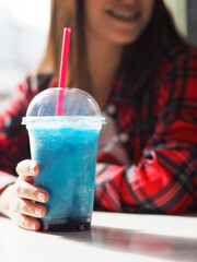 A close-up shot of a refreshing blue slush drink in a clear plastic cup with a dome lid and a red straw. A smiling woman in a red plaid shirt is holding the cup while sitting at a sunlit