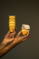 Set of plastic cups for yogurt, puree, jello in hands. Stack of yellow plastic containers for baby food or drinks. Food delivery and takeaway food. Plastic concept. Reusable containers. Copy space.