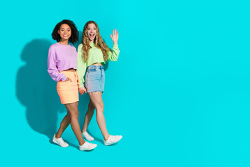 Two stylish young women, smiling and walking together, posing against a bright turquoise background